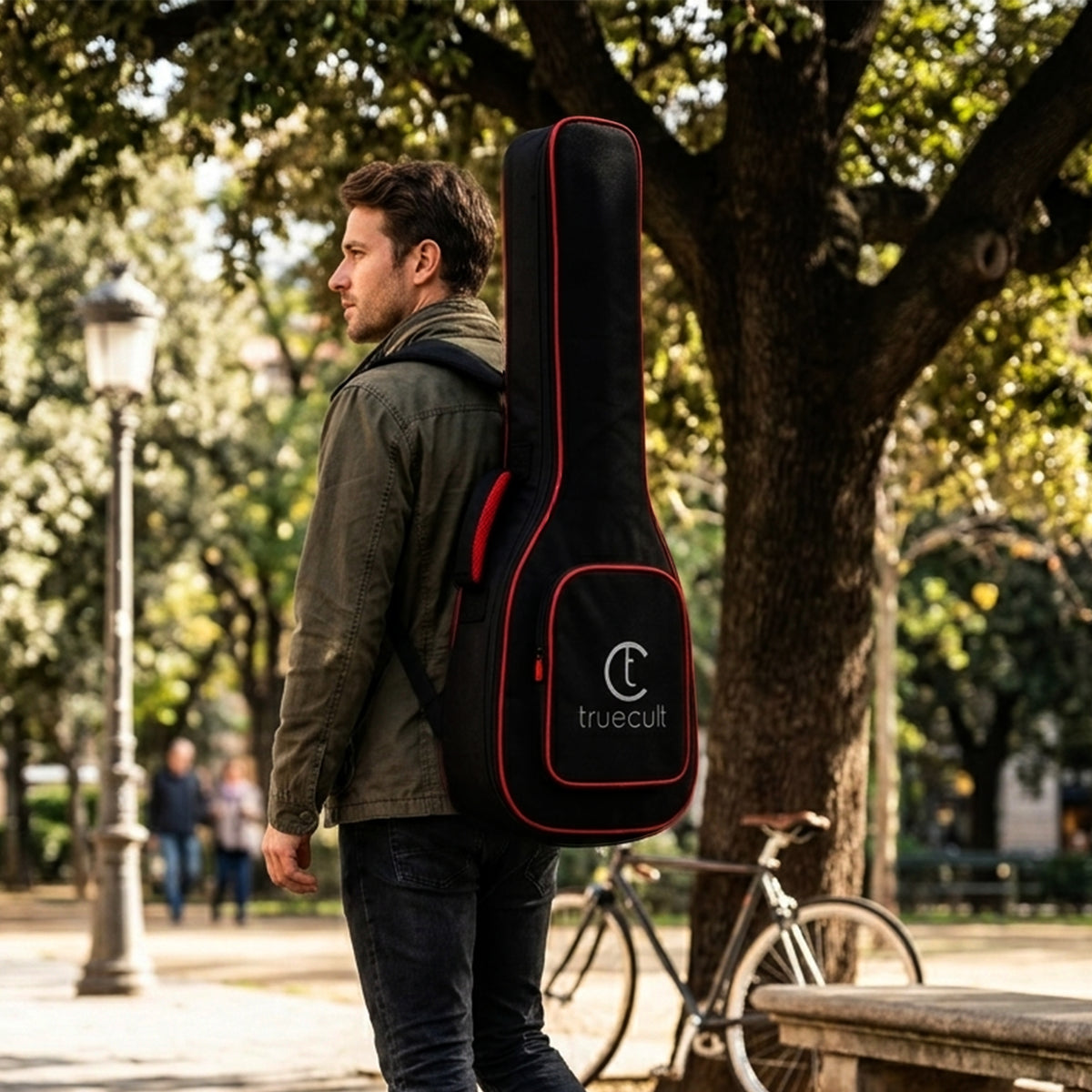 A man wearing a green jacket and dark jeans walks outdoors carrying a black guitar case on his back. The case has red piping and a white 'truecult' logo on the front pocket.